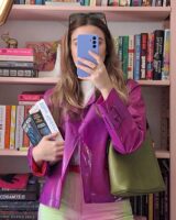 Mulher de pele clara, com cabelo loiro, jaqueta rosa, bolsa verde-escuro. Ela está segurando livros e um celular