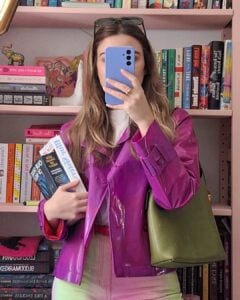 Mulher de pele clara, com cabelo loiro, jaqueta rosa, bolsa verde-escuro. Ela está segurando livros e um celular
