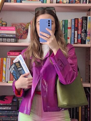 Mulher de pele clara, com cabelo loiro, jaqueta rosa, bolsa verde-escuro. Ela está segurando livros e um celular