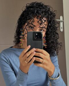 Mulher de pele clara, com blusa azul, brinco colorido e cabelo crespo preto