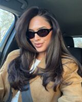 Mulher de pele clara, com blusa branca e casaco caramelo, cabelo escuro escovado e óculos escuros