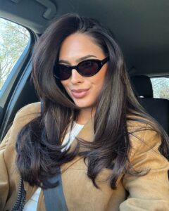 Mulher de pele clara, com blusa branca e casaco caramelo, cabelo escuro escovado e óculos escuros