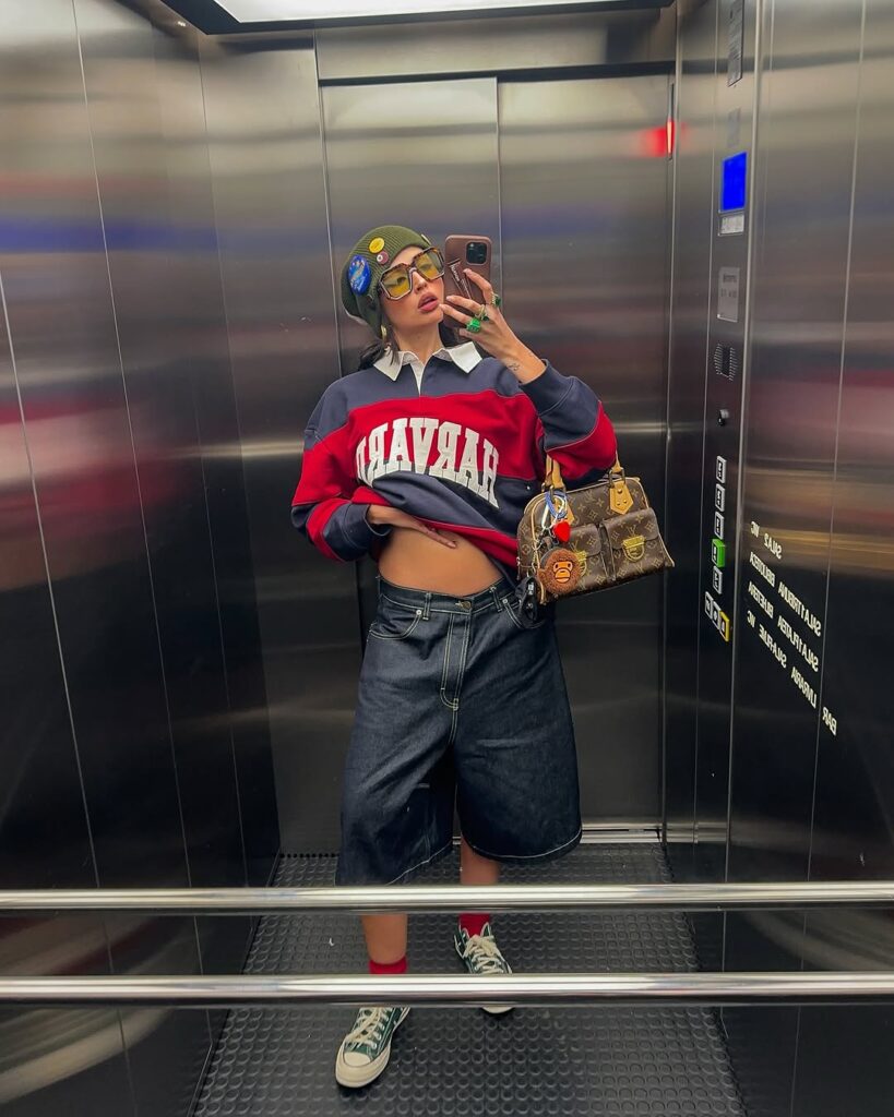 Mulher jovem e magra em foto no elevador. Ela usa suéter listrado e bermuda.