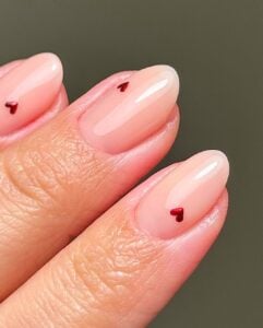 Mão de pele clara, com unha amendoada nude com micro corações em vermelho metálico