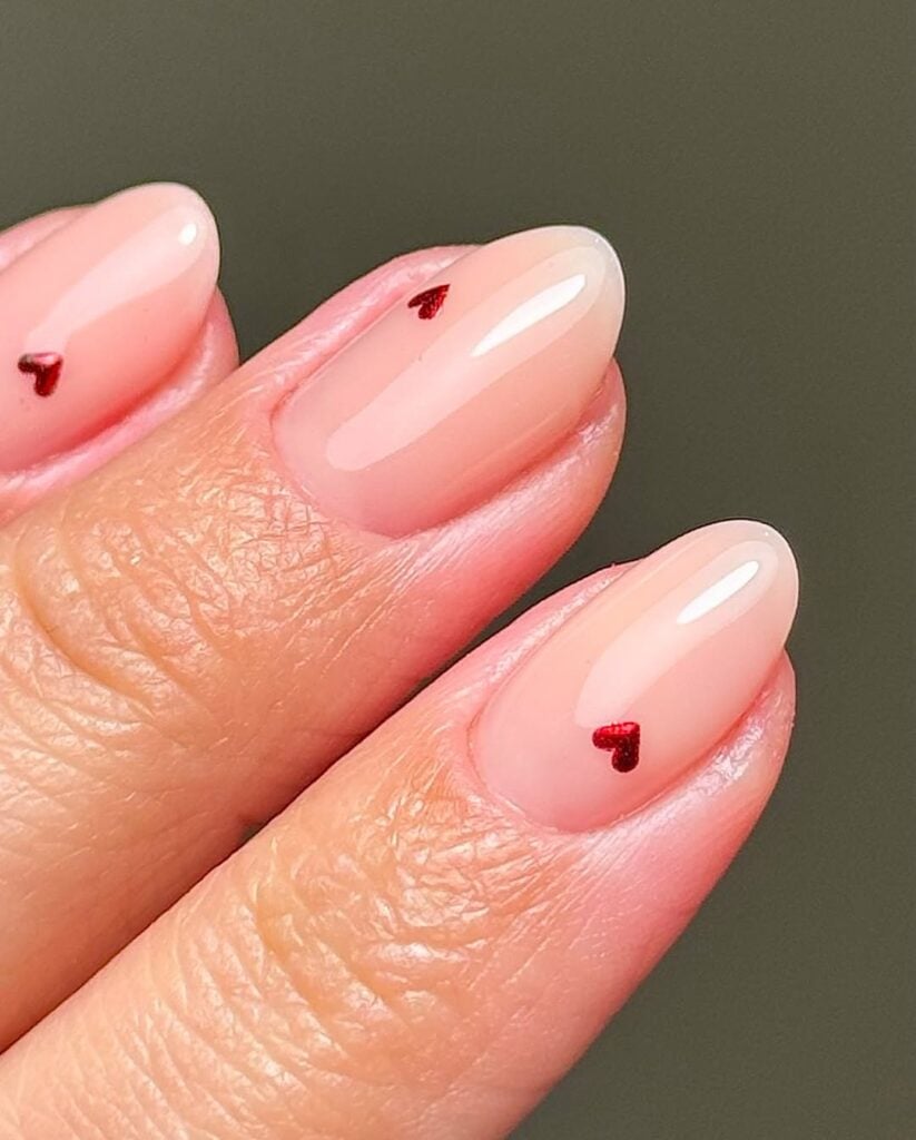 Mão de pele clara, com unha amendoada nude com micro corações em vermelho metálico 