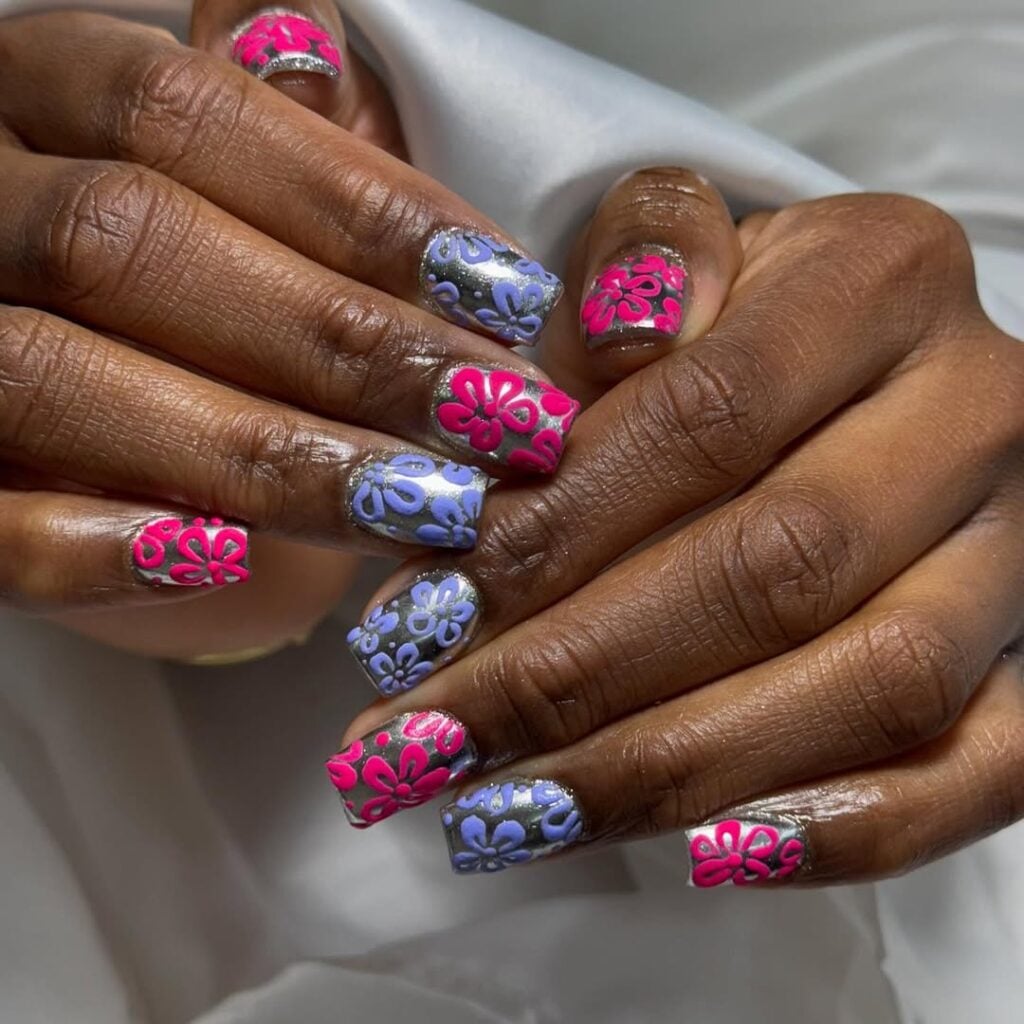 Mãos de pele preta, com unha curta e quadrada metalizada, com flores coloridas