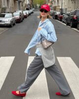 Mulher jovem e magra andando na rua com camisa listrada azul, calça cinza, sapatilhas e boné vermelho e bolsa estampada.