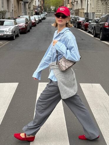 Mulher jovem e magra andando na rua com camisa listrada azul, calça cinza, sapatilhas e boné vermelho e bolsa estampada.