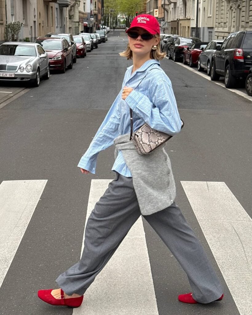 Mulher jovem e magra andando na rua com camisa listrada azul, calça cinza, sapatilhas e boné vermelho e bolsa estampada.