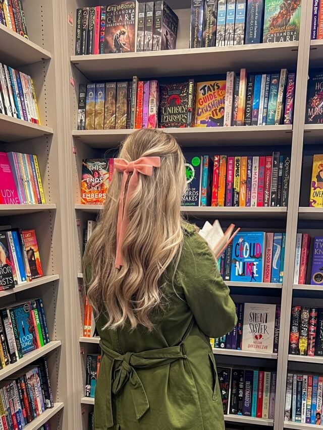 Mulher de pele clara, com casaco verde, cabelo loiro e laço rosa