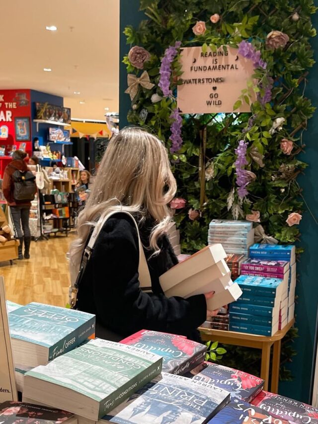 Mulher de pele clara, com cabelo loiro e jaqueta preta. Ela está em uma livraria