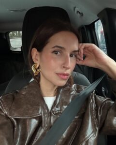 Mulher jovem em selfie dentro do carro. Ela usa jaqueta de couro marrom e brinco dourado grande.