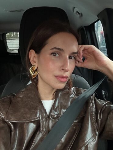 Mulher jovem em selfie dentro do carro. Ela usa jaqueta de couro marrom e brinco dourado grande.