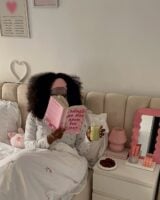 Mulher jovem sentada na cama de pijama. Ela está bebendo chá gelado e lendo um livro.