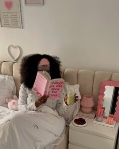 Mulher jovem sentada na cama de pijama. Ela está bebendo chá gelado e lendo um livro.