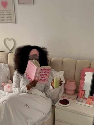 Mulher jovem sentada na cama de pijama. Ela está bebendo chá gelado e lendo um livro.