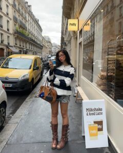 Mulher jovem de pele clara com lookk composto de shorts, bota e suéter listrado
