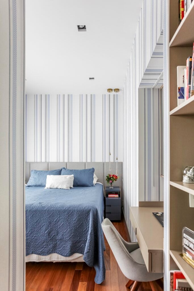 Quarto com teto branco, papel de parede listrado, cabeceira estofada cinza com roupa de cama azul