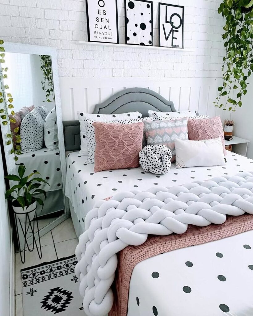 Cama com cabeceira cinza, painel de madeira branco atrás, lençol branco com bolinhas pretas, almofadas em rosa e branco