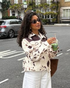 Mulher de pele preta, com cabelo cacheado escuro, calça jeans clara e casaco cow print