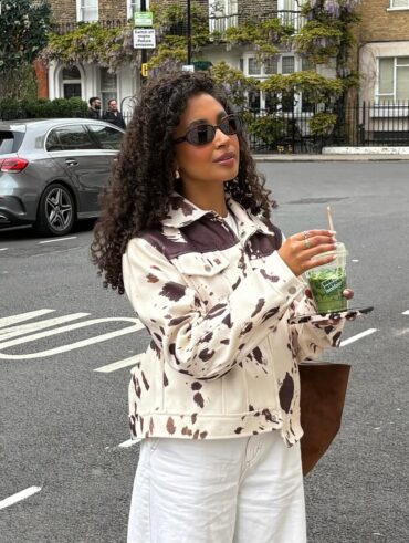 Mulher de pele preta, com cabelo cacheado escuro, calça jeans clara e casaco cow print