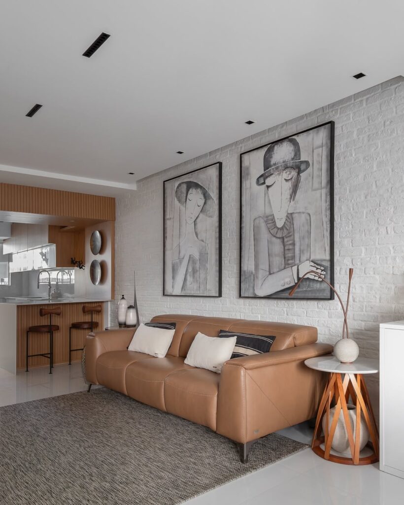 Sala com parede de tijolinhos brancos, quadros em branco e preto e sofá de couro marrom
