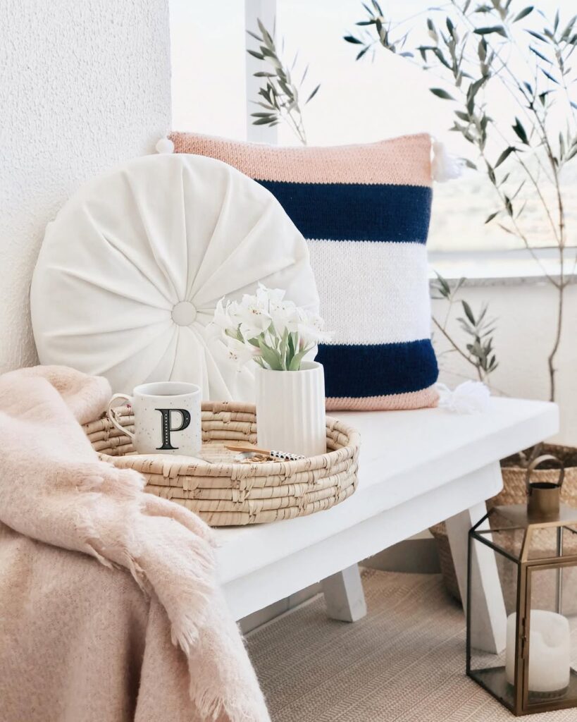Banco de madeira branco com bandeja de palha com caneca e vasinho de flores, e almofadas em branco, rosa e azul-marinho
