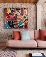 Sala de estar com sofá em estrutura de madeira com estofado rosa-claro, almofadas e quadro colorido em parede de pedras