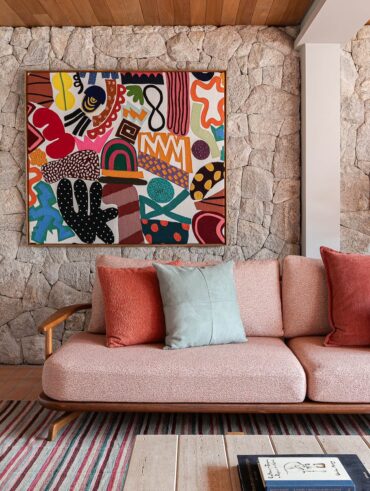 Sala de estar com sofá em estrutura de madeira com estofado rosa-claro, almofadas e quadro colorido em parede de pedras
