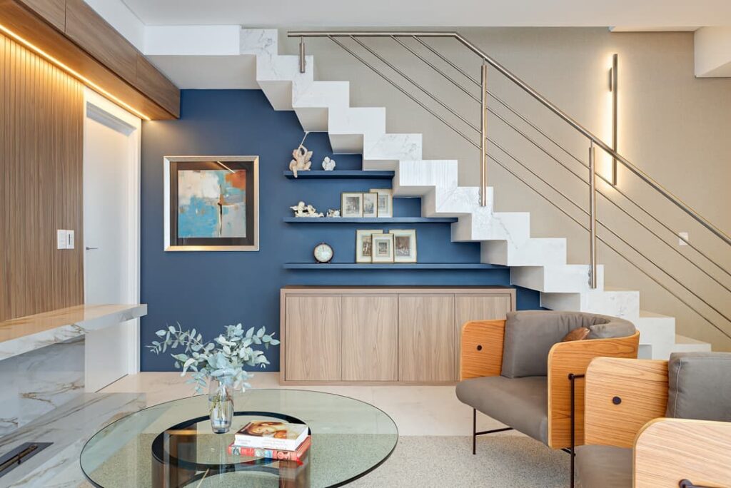 Sala com parede azul com prateleiras debaixo da escada, poltronas com estrutura amadeirada e assento cinza