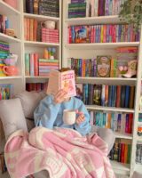 Mulher de pele clara, sentada em uma poltrona com moletom azul-bebê, cobertor rosa. Ela está segurando uma xícara e com o livro aberto no rosto