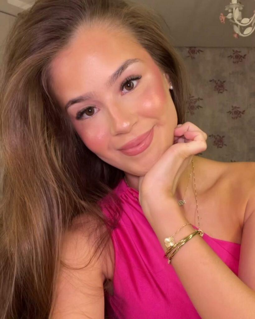 Mulher de pele clara, com vestido rosa, acessórios em dourado, cabelo loiro e maquiagem clean 