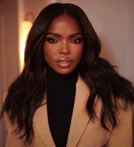 Mulher de pele preta, com blusa preta e blazer bege, cabelo escuro e maquiagem com contorno nos olhos