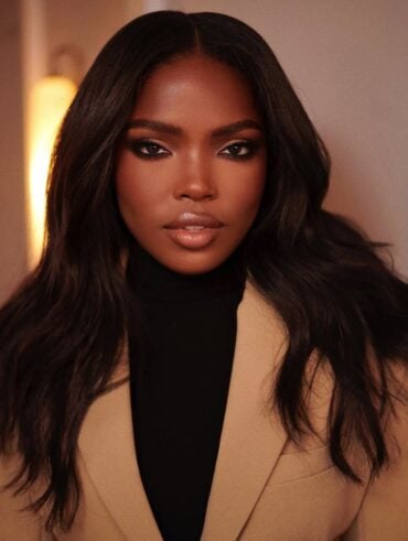 Mulher de pele preta, com blusa preta e blazer bege, cabelo escuro e maquiagem com contorno nos olhos