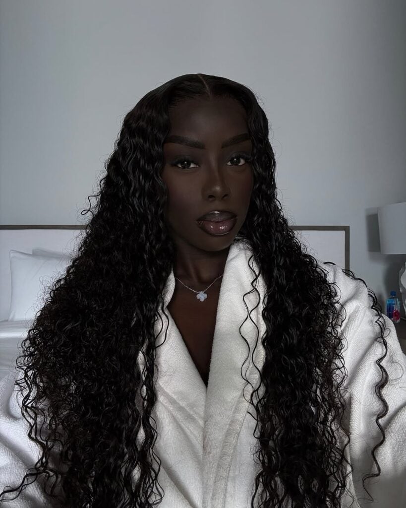 Mulher de pele preta, com roupão branco, acessórios em prata, cabelo cacheado escuro e maquiagem clean 