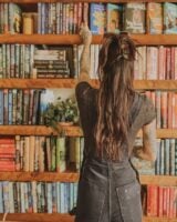 Mulher de pele clara, com blusa e jardineira preta, cabelo castanho preso em coque. Ela está na frente de uma estante de livros