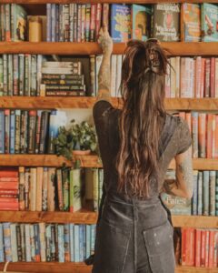 Mulher de pele clara, com blusa e jardineira preta, cabelo castanho preso em coque. Ela está na frente de uma estante de livros