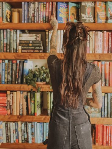 Mulher de pele clara, com blusa e jardineira preta, cabelo castanho preso em coque. Ela está na frente de uma estante de livros
