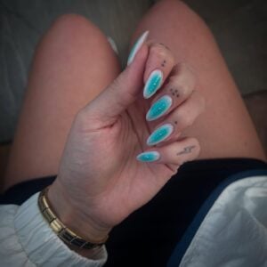 Mão de pele clara, com acessórios em dourado e unha amendoada aura com esmalte azul e branco