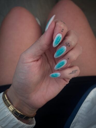 Mão de pele clara, com acessórios em dourado e unha amendoada aura com esmalte azul e branco