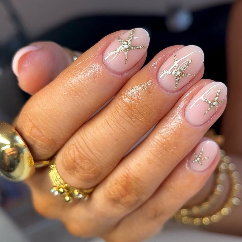 Mão de pele clara, com acessórios em dourado e unha amendoada nude com estrela dourada