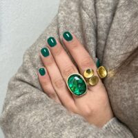 Mão de pele clara, com anéis em verde e dourado e unha curta e arredondada verde-esmeralda