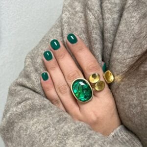 Mão de pele clara, com anéis em verde e dourado e unha curta e arredondada verde-esmeralda