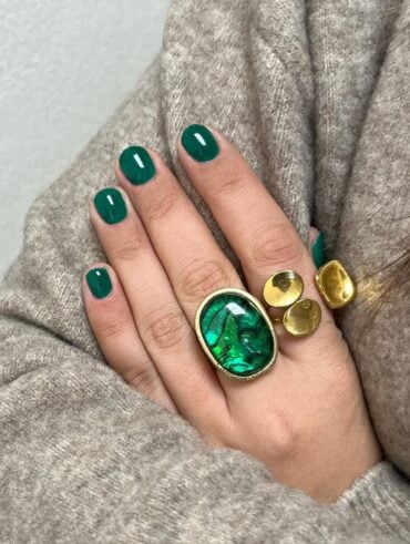 Mão de pele clara, com anéis em verde e dourado e unha curta e arredondada verde-esmeralda