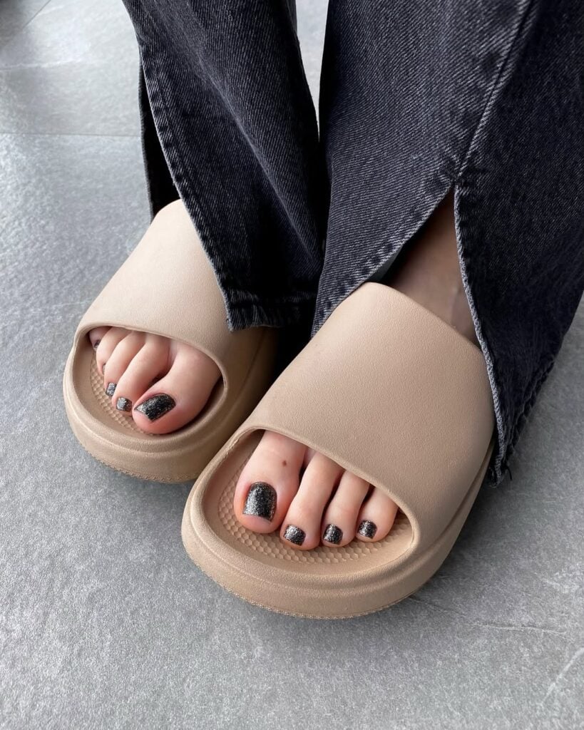 Pés de pele clara, com chinelo slide e pedicure com esmalte preto brilhoso