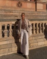 Mulher jovem com look com calça jeans branca, cinto marrom e casaco marrom claro