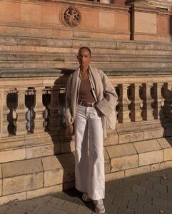 Mulher jovem com look com calça jeans branca, cinto marrom e casaco marrom claro