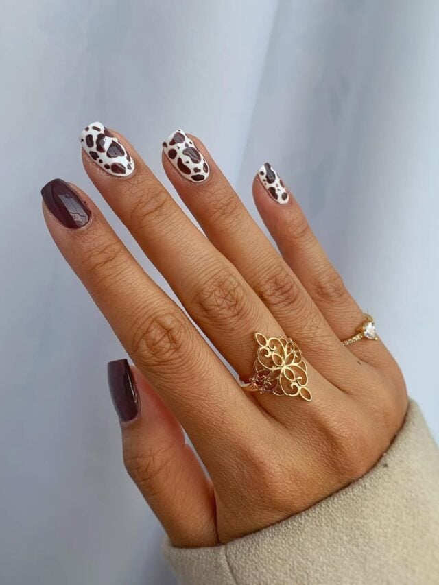 Mão de pele clara, com anéis em dourado e unha quadrada marrom com animal print de vaquinha