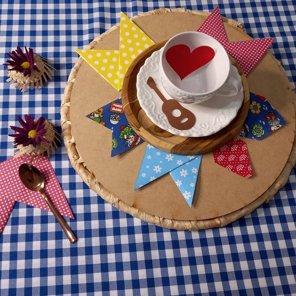 Mesa com toalha xadrez, sousplat de palha e papelão e bandeirinhas e balões coloridos de papel