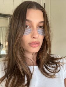 Hailey Bieber, com blusa branca, cabelo castanho e patches na área dos olhos